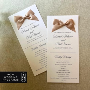 May include: Two ivory wedding programs with a peach satin bow on the top of each. The programs list the wedding ceremony order, including the names of the bride and groom, Brandi Johnson and Brell Vescovi, and the date and location of the wedding. The text reads "Welcome to the wedding of Brandi Johnson and Brell Vescovi. Saturday, March 22nd, 2025. Fuquay-Varina, North Carolina."