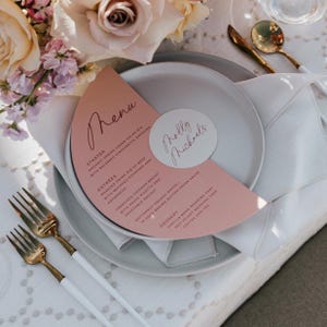 May include: A semi-circular dusty pink menu card with black text and a circular white name tag reading Molly Michaels, placed on a light gray plate. The menu lists starter, entree, and dessert options, accompanied by gold-toned cutlery.