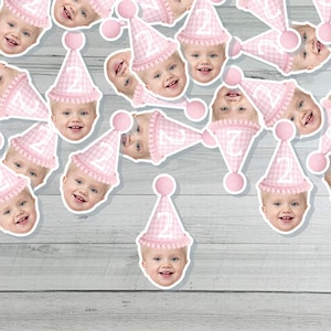 May include: A collection of stickers featuring a baby's face and a pink gingham party hat with the number 2. The stickers are scattered across a light gray wooden surface, creating a festive and celebratory theme.