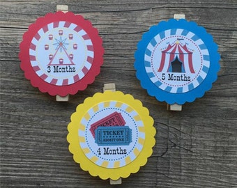 Puede incluir: Tres círculos de papel coloridos con bordes festoneados y pinzas para la ropa adjuntas. Cada círculo presenta una imagen diferente con temática de circo: una noria, una carpa y una entrada. Los círculos están etiquetados con los números "3 meses", "4 meses" y "5 meses".