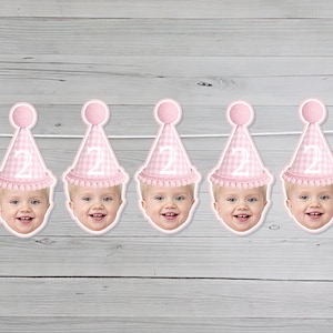 May include: A festive banner featuring a series of smiling baby faces wearing pink party hats with the number "2" on them. The hats have a pink and white checkered pattern. The banner is strung against a light gray wooden background, with clear balloons on the sides.