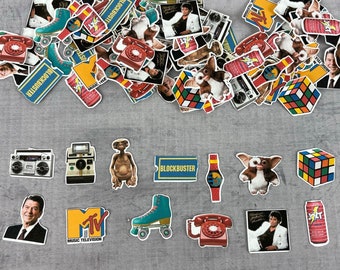 May include: A collection of colorful stickers featuring various 80s and 90s pop culture icons, including ET, the '80s movie character, a boombox, a Rubik's Cube, and the MTV logo. The stickers are arranged on a gray surface.