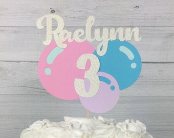 Puede incluir: Un pastel blanco con glaseado blanco de crema de mantequilla y un adorno de pastel de purpurina plateada con el nombre "Raelynn" y el número "3" en purpurina plateada. El adorno de pastel presenta tres globos en rosa, azul y morado.