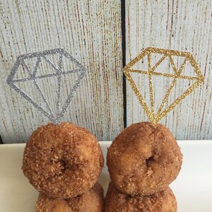 May include: Two stacks of mini donuts topped with silver and gold glitter diamond cupcake toppers. The donuts are on a white rectangular plate.