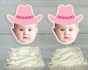 May include: Two pink cowboy hats with white polka dots and a baby's face in the center of each hat. The hats are on top of cupcakes with white frosting.