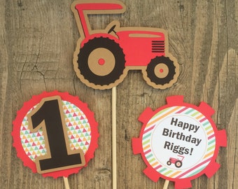 May include: Three red and brown paper cupcake toppers with a red tractor, a number one, and a gear with the text "Happy Birthday Riggs!" and a small tractor image.