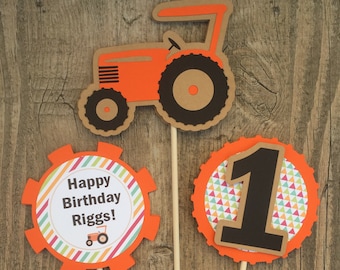 May include: Orange and brown tractor and number one cake toppers with a personalized "Happy Birthday Riggs!" message. The toppers are on wooden sticks, perfect for a first birthday celebration with a farm theme.