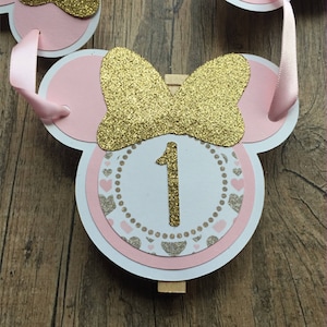 Op de afbeelding: Roze en gouden Minnie Mouse-themabanner met een witte cirkel met het nummer 1 in gouden glitter. De banner is aan elkaar geregen met roze lint en heeft een houten wasknijper.