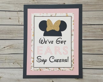 May include: A pink and gold printable sign with a black silhouette of Mickey Mouse's ears with a gold bow. The text reads "We've Got Ears Say Cheers!"