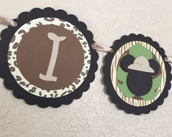 Può includere: Uno striscione nero e marrone a tema giungla. Lo striscione ha le lettere "I" e "A" e un'immagine di un animale da safari con un cappello.