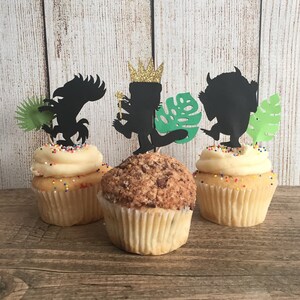 May include: Three cupcakes decorated with Where the Wild Things Are themed toppers. The toppers are black silhouettes of the characters, including a monster with a crown, a wild thing, and a monster with leaves. The cupcakes are frosted with vanilla frosting and colorful sprinkles.