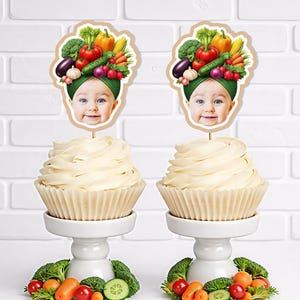 May include: Two cupcakes with white frosting, each topped with a baby face wearing a vegetable crown. The cupcakes sit on white pedestals, surrounded by fresh vegetables like broccoli, carrots, and cucumbers. The background is a white brick wall.