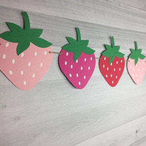 May include: A string of paper strawberry cutouts in pink, red, and light pink with white polka dots. The strawberries are strung together with twine and are hanging against a wood background.