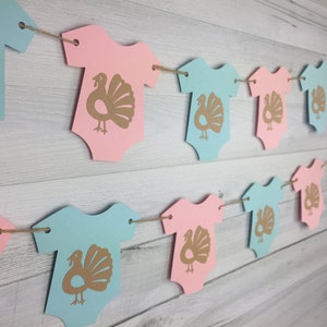 May include: A garland of pink and blue baby onesies with gold turkey silhouettes. The onesies are strung together with twine.