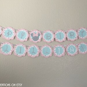 May include: A pink and blue banner with silver glitter letters that reads "Happy Birthday Lillian". The banner has a scalloped edge and features small silver glitter Minnie Mouse heads.