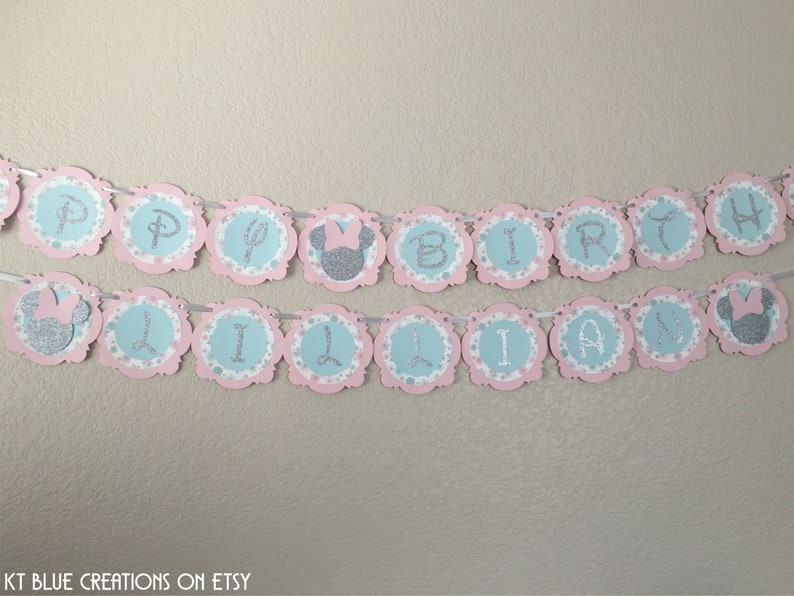 May include: A pink and blue banner with silver glitter letters that reads "Happy Birthday Lillian". The banner has a scalloped edge and features small silver glitter Minnie Mouse heads.