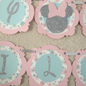 May include: A pink and blue banner featuring a silver glitter Minnie Mouse head and the letters "One" in silver glitter. The banner is decorated with snowflakes and floral patterns.