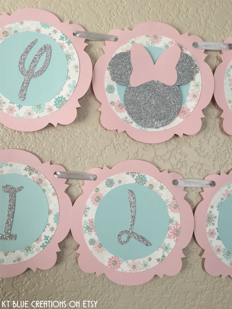 May include: A pink and blue banner featuring a silver glitter Minnie Mouse head and the letters "One" in silver glitter. The banner is decorated with snowflakes and floral patterns.