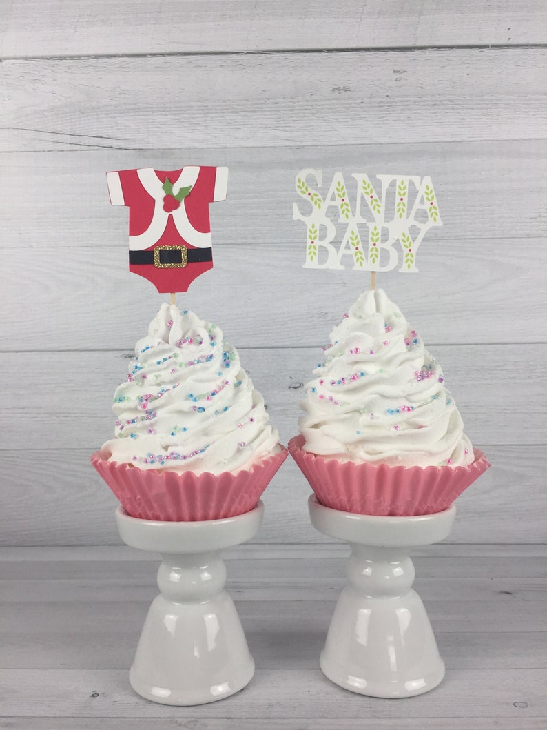 K&ouml;nnte beinhalten: Zwei wei&szlig;e Cupcakes mit rosa Cupcake-F&ouml;rmchen und wei&szlig;em Frosting, der mit bunten Streuseln verziert ist. Die Cupcakes stehen auf wei&szlig;en Kuchenst&auml;ndern. Ein Cupcake hat einen Santa-Baby-Topper und der andere einen Santa-Outfit-Topper.
