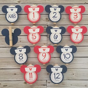 May include: Twelve red and blue Mickey Mouse themed monthly milestone cards with a sailor hat and anchor design. The cards are numbered from NB to 12 months.