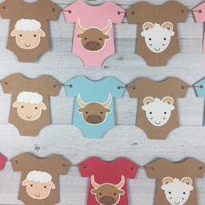May include: A brown, pink, blue, and red baby onesie banner with alternating images of a white lamb and a brown bull. The banner is strung together with twine.