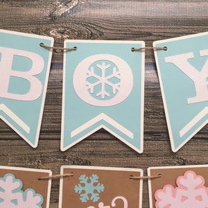 May include: A banner that spells out "BOY" in white letters on light blue pennants, with snowflake accents. The banner is strung on twine and set against a wooden background. Additional pennants below feature snowflakes.