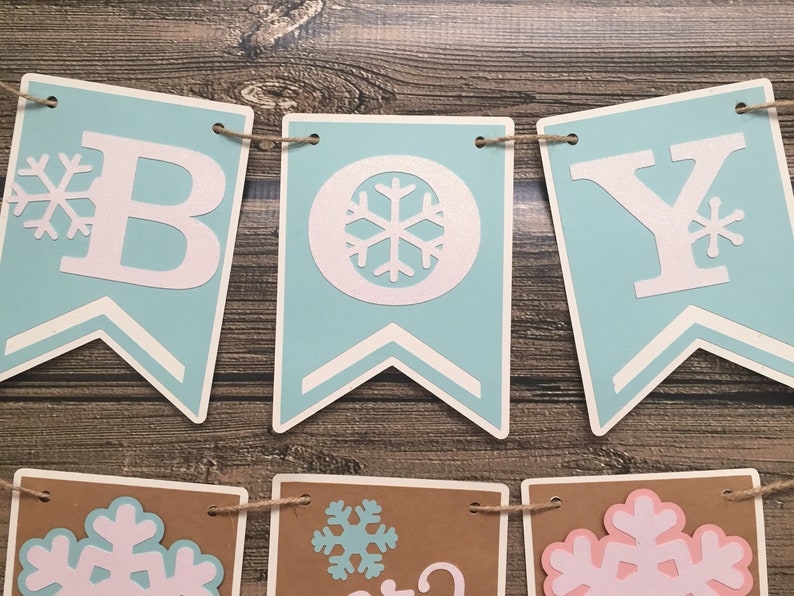 May include: A banner that spells out "BOY" in white letters on light blue pennants, with snowflake accents. The banner is strung on twine and set against a wooden background. Additional pennants below feature snowflakes.
