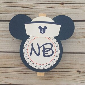 May include: A blue and white nautical themed Mickey Mouse head with a sailor hat and a circle with anchors and the initials "NB" in the center.