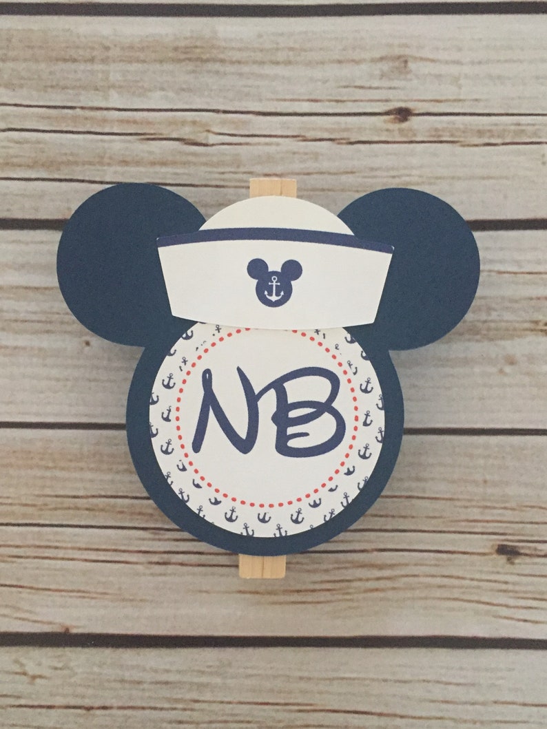 May include: A blue and white nautical themed Mickey Mouse head with a sailor hat and a circle with anchors and the initials "NB" in the center.