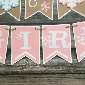 May include: A banner spelling "GIRL" in white letters on pink pennants, with snowflake accents. Additional pennants with snowflakes and the word "GIRL" are visible. The banner is strung on twine, with a wooden background.