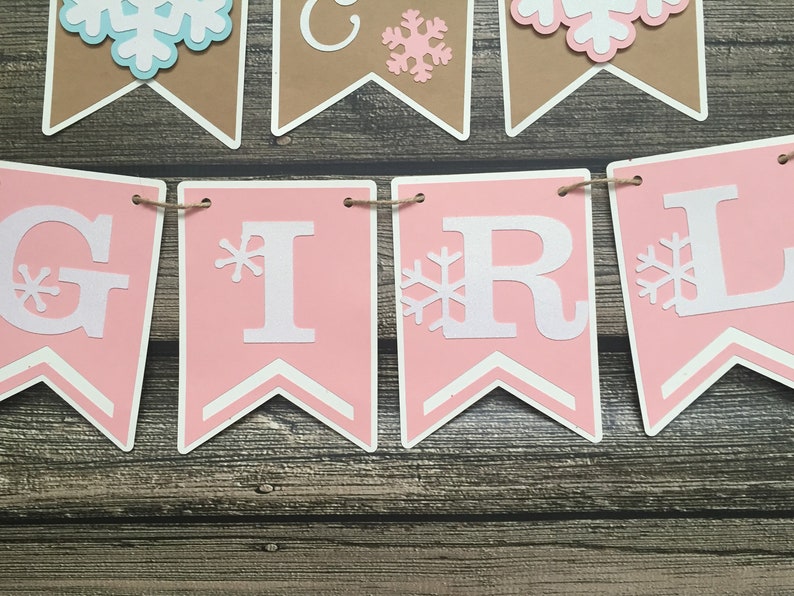 May include: A banner spelling "GIRL" in white letters on pink pennants, with snowflake accents. Additional pennants with snowflakes and the word "GIRL" are visible. The banner is strung on twine, with a wooden background.
