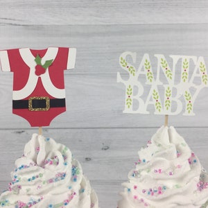 K&ouml;nnte beinhalten: Zwei Cupcakes, verziert mit wei&szlig;em Frosting und Streuseln. Ein Cupcake hat einen roten und wei&szlig;en Weihnachtsmann-Anzug-Topper, der andere einen wei&szlig;en Topper mit dem Text "SANTA BABY" in gr&uuml;nen Buchstaben mit Stechpalmen-Akzenten.