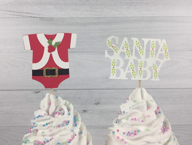 K&ouml;nnte beinhalten: Zwei Cupcakes, verziert mit wei&szlig;em Frosting und Streuseln. Ein Cupcake hat einen roten und wei&szlig;en Weihnachtsmann-Anzug-Topper, der andere einen wei&szlig;en Topper mit dem Text "SANTA BABY" in gr&uuml;nen Buchstaben mit Stechpalmen-Akzenten.