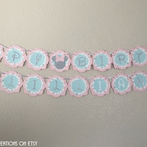 May include: A pink and blue birthday banner with silver glitter letters that reads "Happy Birthday Lillian". The banner has a scalloped edge and features Minnie Mouse ears with a pink bow.