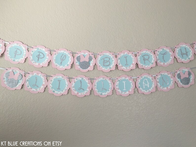 May include: A pink and blue birthday banner with silver glitter letters that reads "Happy Birthday Lillian". The banner has a scalloped edge and features Minnie Mouse ears with a pink bow.