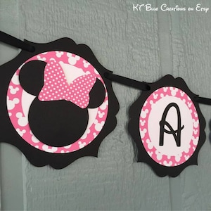 Op de afbeelding: Zwarte en roze Minnie Mouse-banner met de letter "A" in het midden van een van de cirkels. De banner is versierd met witte stippen en Minnie Mouse-oren.