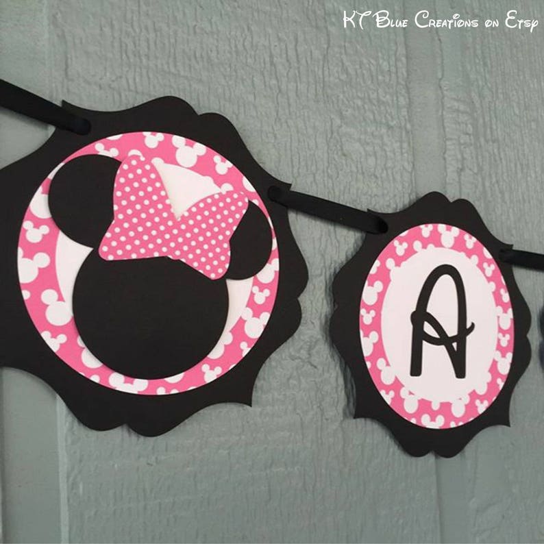 Op de afbeelding: Zwarte en roze Minnie Mouse-banner met de letter "A" in het midden van een van de cirkels. De banner is versierd met witte stippen en Minnie Mouse-oren.