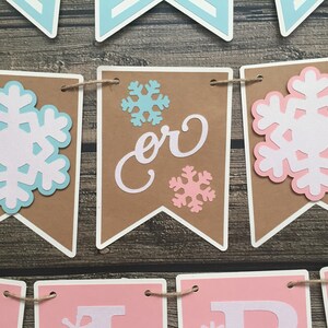 May include: A banner with brown pennants, white borders, and snowflake cutouts in light blue, white, and pink. The word "or" is displayed on one pennant. The banner is strung with twine.