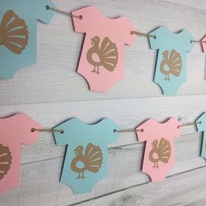 May include: A gender neutral baby shower banner with alternating pink and blue onesies with gold turkey silhouettes. The banner is strung with twine.