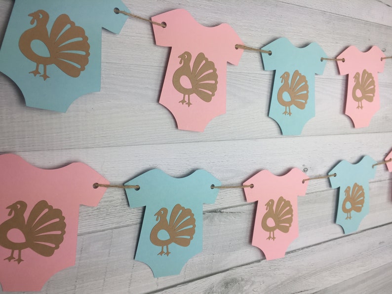 May include: A gender neutral baby shower banner with alternating pink and blue onesies with gold turkey silhouettes. The banner is strung with twine.