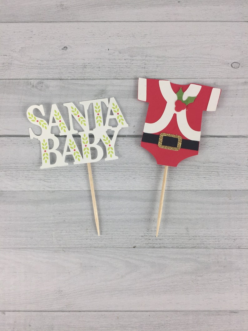 K&ouml;nnte beinhalten: Zwei Tortendekorationen, eine mit den Worten "Santa Baby" in Wei&szlig; mit gr&uuml;nen Stechpalmen-Akzenten und die andere ein rotes Weihnachtsmannkost&uuml;m mit wei&szlig;em Besatz und einem goldenen G&uuml;rtel.