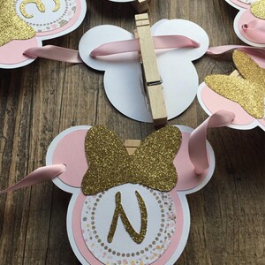 Op de afbeelding: Roze en gouden Minnie Mouse-themabanner met een letter N en een strik. De banner is gemaakt van karton en heeft een glitterafwerking.