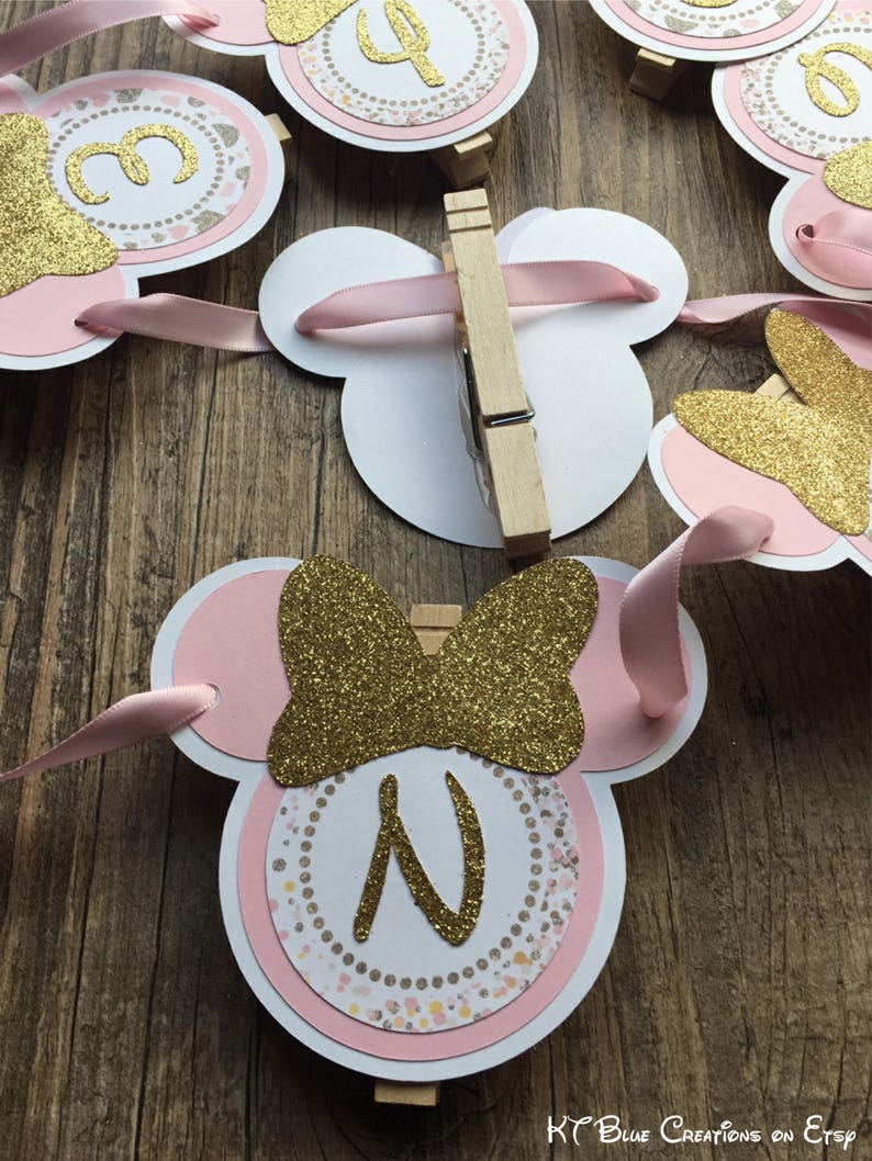 Op de afbeelding: Roze en gouden Minnie Mouse-themabanner met een letter N en een strik. De banner is gemaakt van karton en heeft een glitterafwerking.
