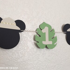 Puede incluir: Una pancarta de Mickey Mouse en blanco y negro con hojas verdes y el n&uacute;mero uno. La pancarta es perfecta para una fiesta de primer cumplea&ntilde;os.
