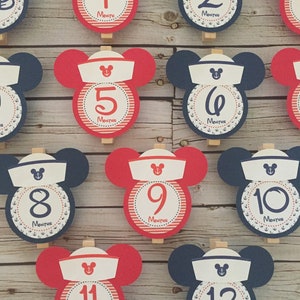 May include: Twelve red and blue Mickey Mouse themed monthly milestone cards with white sailor hats and anchors. The cards are numbered 1 through 12.