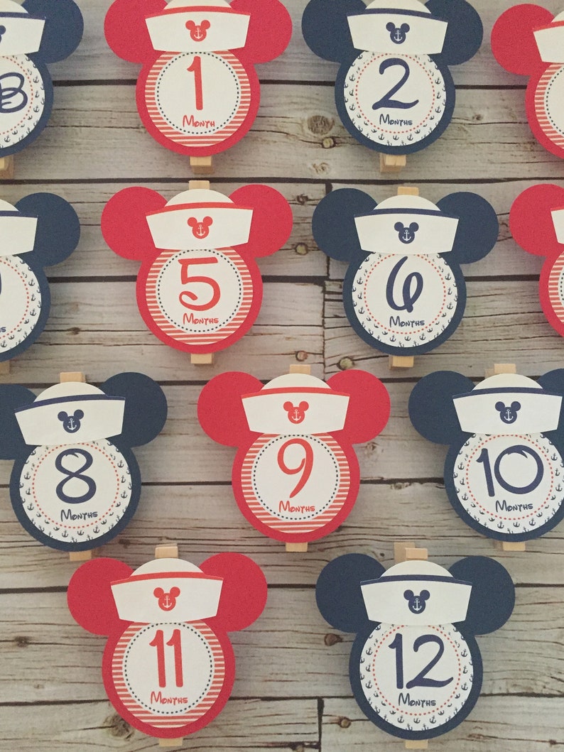May include: Twelve red and blue Mickey Mouse themed monthly milestone cards with white sailor hats and anchors. The cards are numbered 1 through 12.