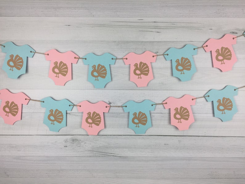 May include: A gender neutral baby shower banner with alternating pink and blue baby onesies with a gold turkey graphic on each onesie.