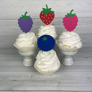 Peut inclure: Quatre cupcakes avec un gla&ccedil;age blanc et des garnitures de fruits diff&eacute;rentes. Les garnitures sont une fraise rouge, une m&ucirc;re violette, une framboise rose et une myrtille bleue.