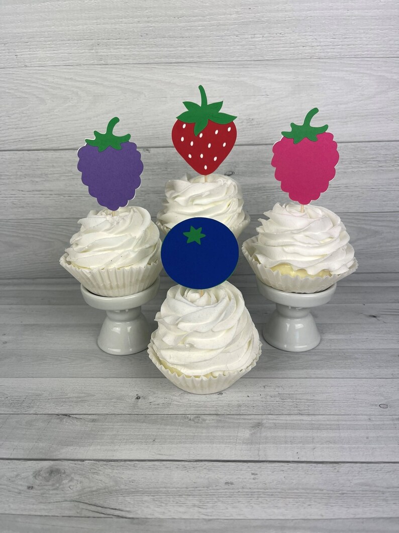 Peut inclure: Quatre cupcakes avec un gla&ccedil;age blanc et des garnitures de fruits diff&eacute;rentes. Les garnitures sont une fraise rouge, une m&ucirc;re violette, une framboise rose et une myrtille bleue.