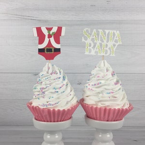 K&ouml;nnte beinhalten: Zwei rosa Cupcake-F&ouml;rmchen mit wei&szlig;em Frosting und bunten Streuseln. Ein Cupcake hat einen &bdquo;Santa Baby&ldquo;-Topper und der andere einen Santa-Baby-Strampler-Topper.
